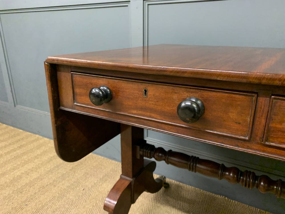 A George IV mahogany sofa table featuring two drawers with dark wooden knobs, drop-leaf side flaps, a turned central stretcher, and splayed legs on brass castors. View IV