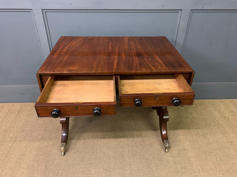 A George IV mahogany sofa table featuring two drawers with dark wooden knobs, drop-leaf side flaps, a turned central stretcher, and splayed legs on brass castors. View V