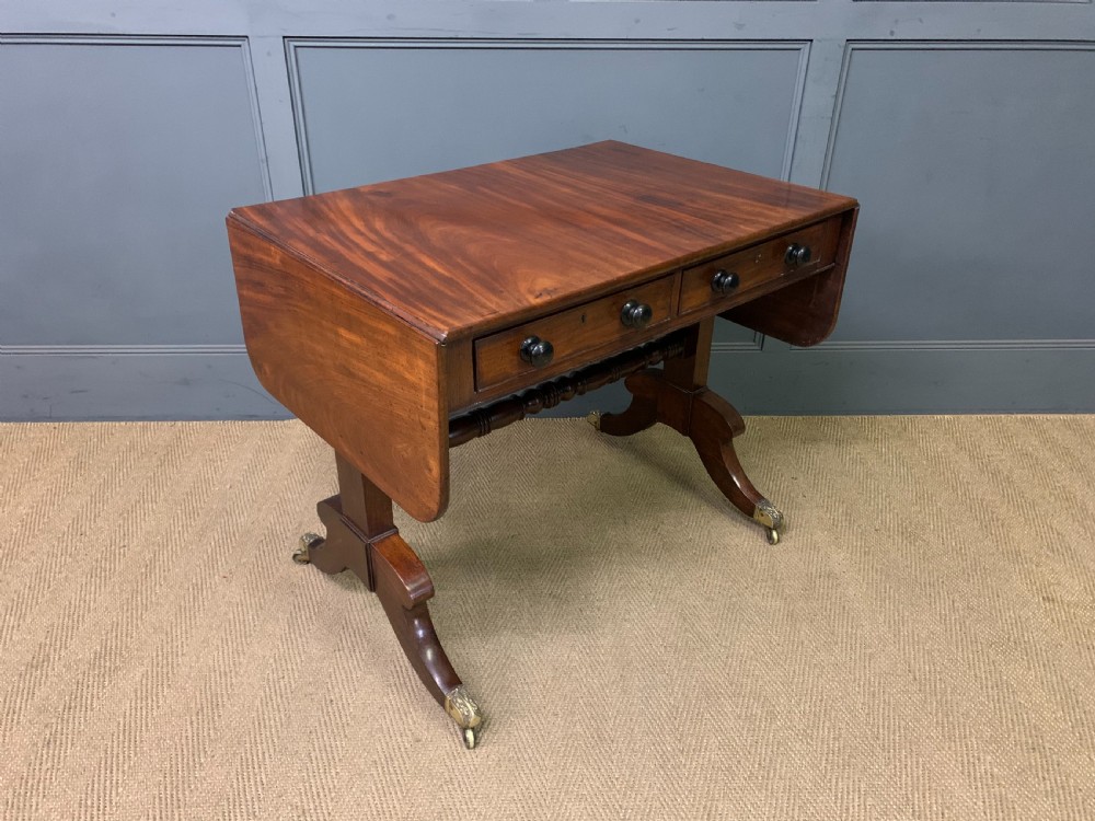 A George IV mahogany sofa table featuring two drawers with dark wooden knobs, drop-leaf side flaps, a turned central stretcher, and splayed legs on brass castors. View VI