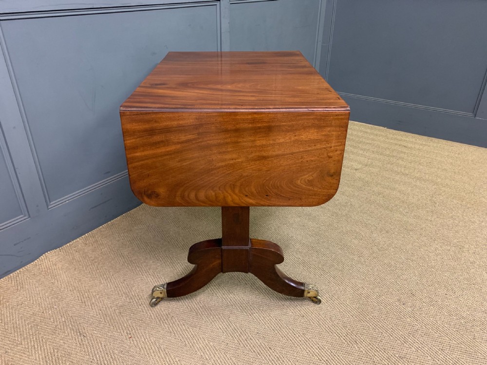 A George IV mahogany sofa table featuring two drawers with dark wooden knobs, drop-leaf side flaps, a turned central stretcher, and splayed legs on brass castors. View VII