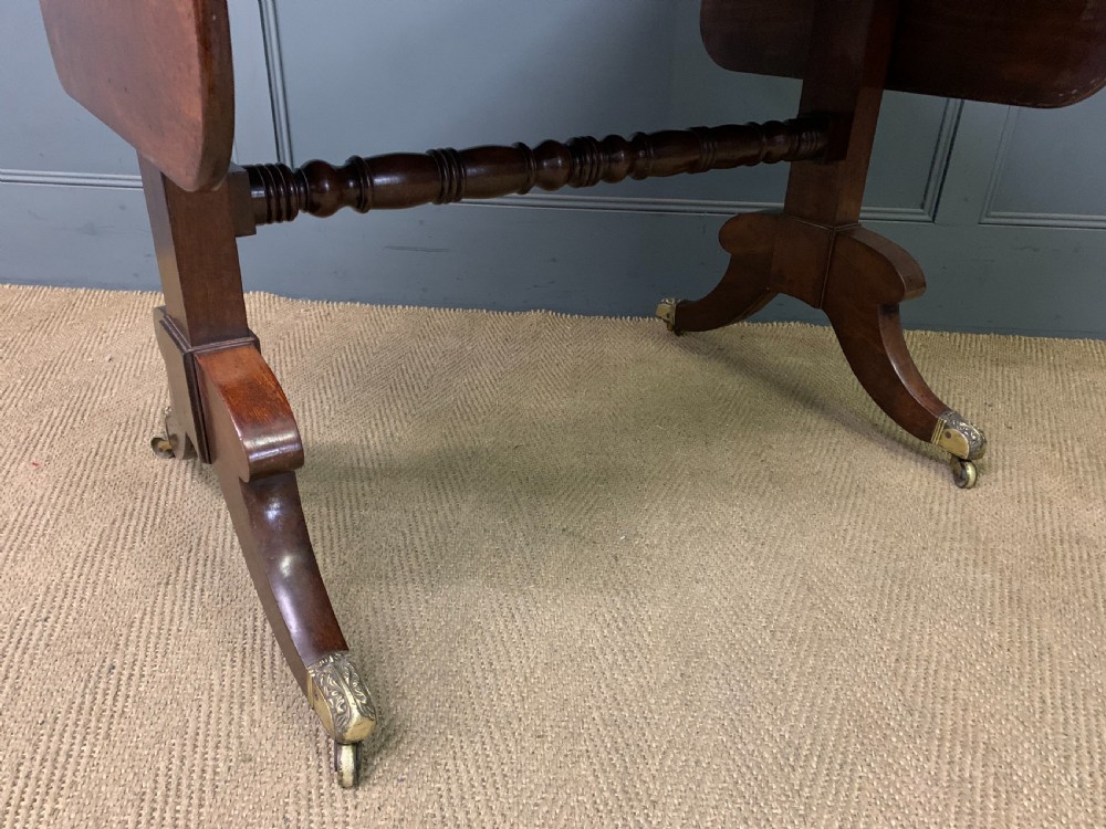 A George IV mahogany sofa table featuring two drawers with dark wooden knobs, drop-leaf side flaps, a turned central stretcher, and splayed legs on brass castors. View IX