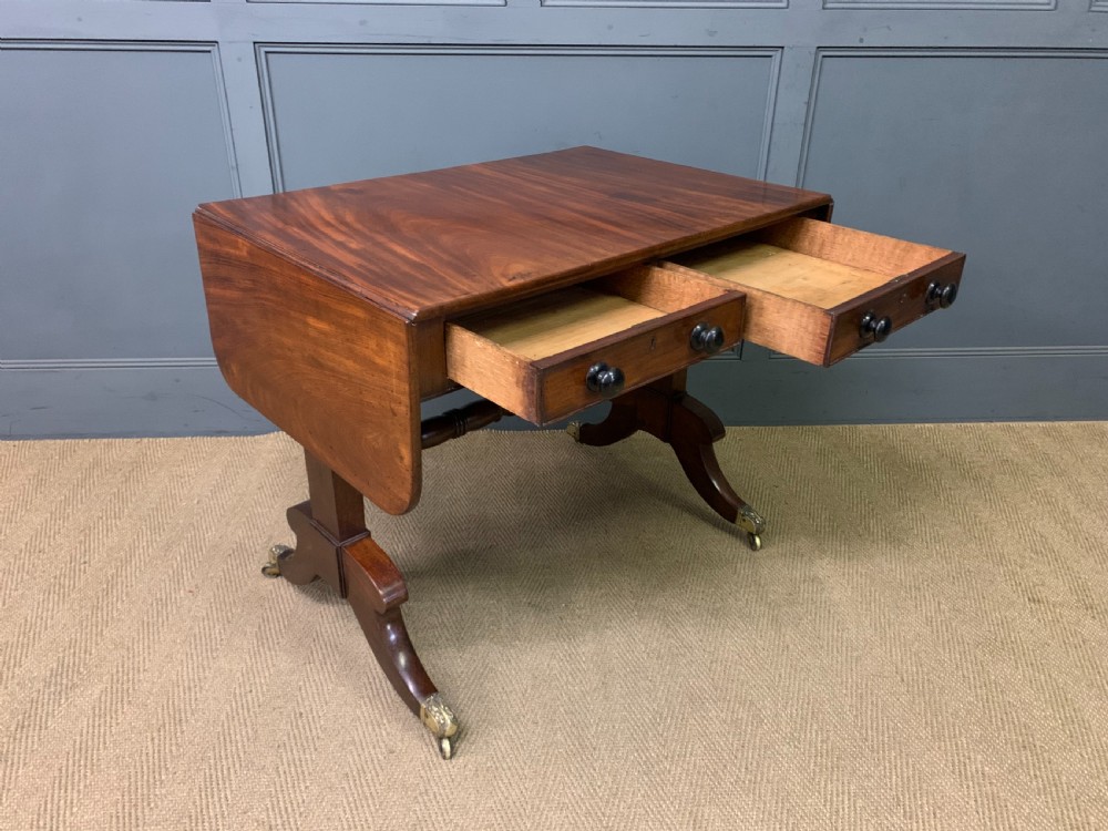 A George IV mahogany sofa table featuring two drawers with dark wooden knobs, drop-leaf side flaps, a turned central stretcher, and splayed legs on brass castors. View X