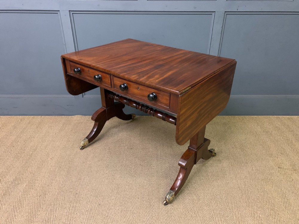 A George IV mahogany sofa table featuring two drawers with dark wooden knobs, drop-leaf side flaps, a turned central stretcher, and splayed legs on brass castors. View XII