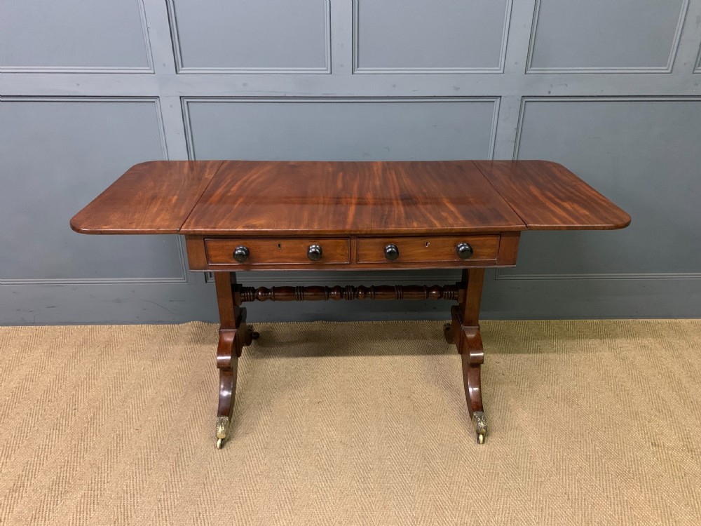 A George IV mahogany sofa table featuring two drawers with dark wooden knobs, drop-leaf side flaps, a turned central stretcher, and splayed legs on brass castors. View XIII