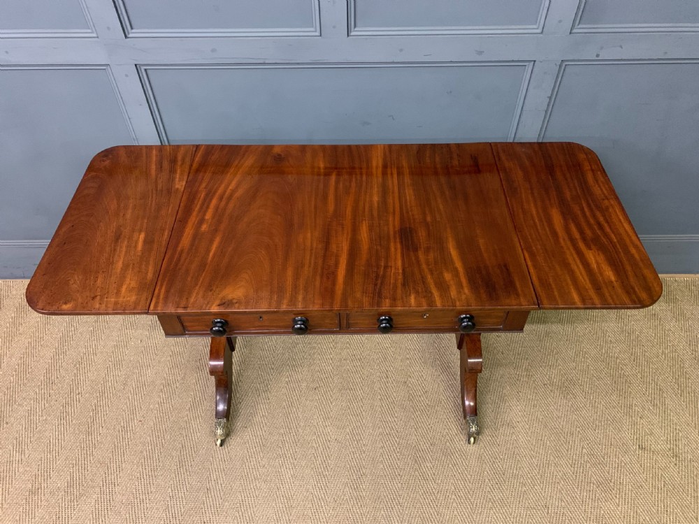 A George IV mahogany sofa table featuring two drawers with dark wooden knobs, drop-leaf side flaps, a turned central stretcher, and splayed legs on brass castors. View XIV