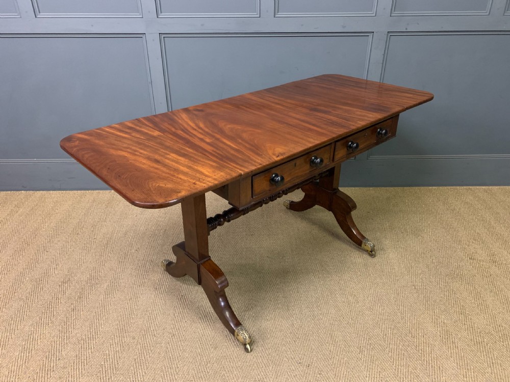A George IV mahogany sofa table featuring two drawers with dark wooden knobs, drop-leaf side flaps, a turned central stretcher, and splayed legs on brass castors. View XV