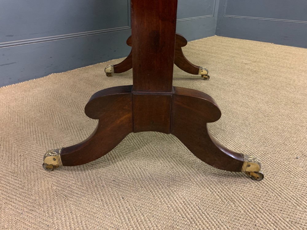 A George IV mahogany sofa table featuring two drawers with dark wooden knobs, drop-leaf side flaps, a turned central stretcher, and splayed legs on brass castors. View XVII
