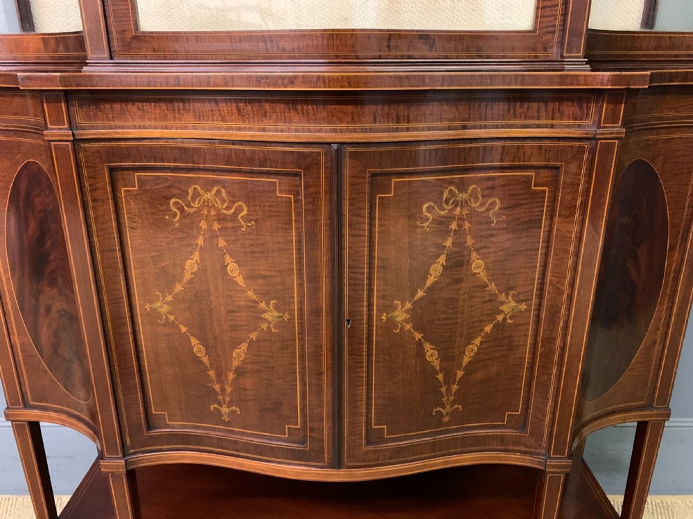 An Edwardian inlaid mahogany display cabinet featuring a glazed upper section, bow-front lower doors with marquetry panels, and tapered legs with a lower shelf. View V