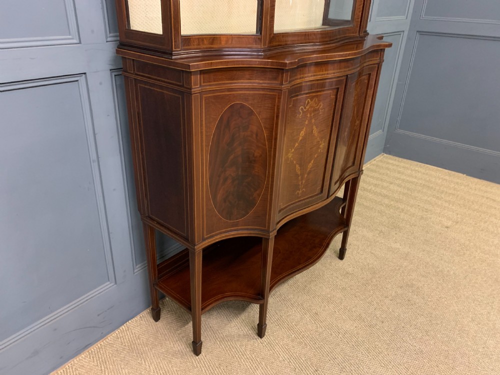 An Edwardian inlaid mahogany display cabinet featuring a glazed upper section, bow-front lower doors with marquetry panels, and tapered legs with a lower shelf. View VI
