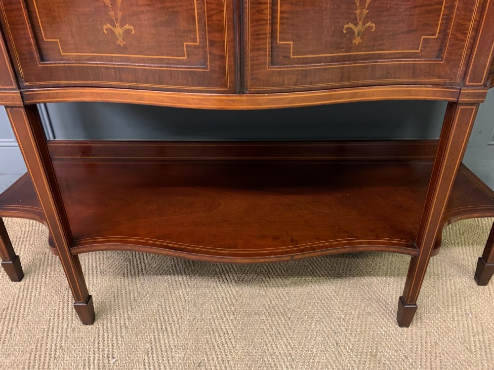 An Edwardian inlaid mahogany display cabinet featuring a glazed upper section, bow-front lower doors with marquetry panels, and tapered legs with a lower shelf. View X