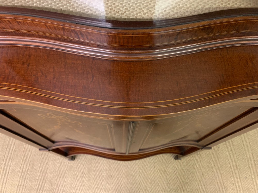 An Edwardian inlaid mahogany display cabinet featuring a glazed upper section, bow-front lower doors with marquetry panels, and tapered legs with a lower shelf. View XVII