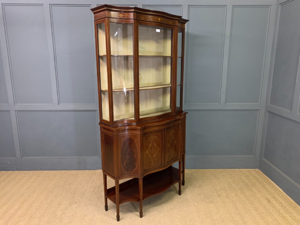 An Edwardian inlaid mahogany display cabinet featuring a glazed upper section, bow-front lower doors with marquetry panels, and tapered legs with a lower shelf. View XXVI