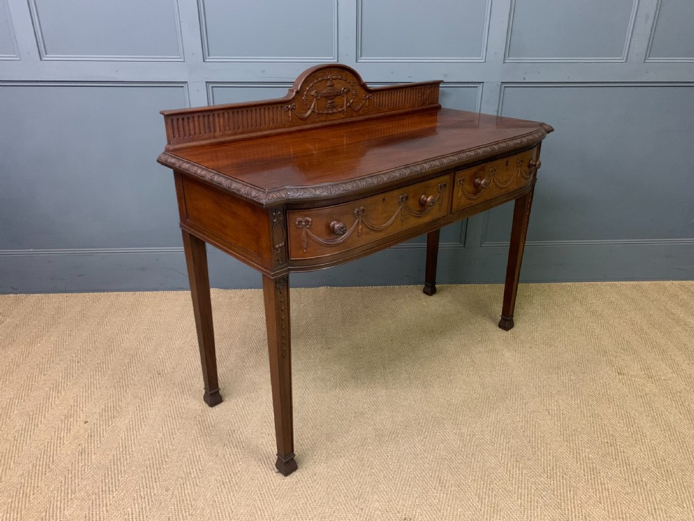 An Adam style mahogany side table featuring a carved upstand with an urn motif, relief drapery swags on the drawer fronts, and square fluted legs.