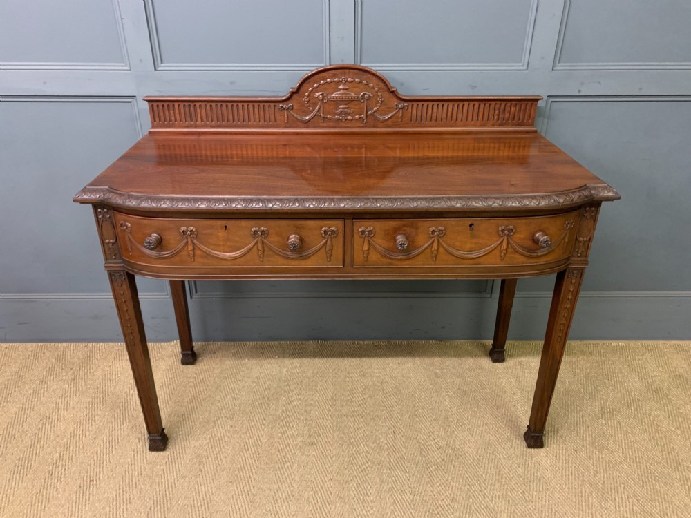 An Adam style mahogany side table featuring a carved upstand with an urn motif, relief drapery swags on the drawer fronts, and square fluted legs. View II
