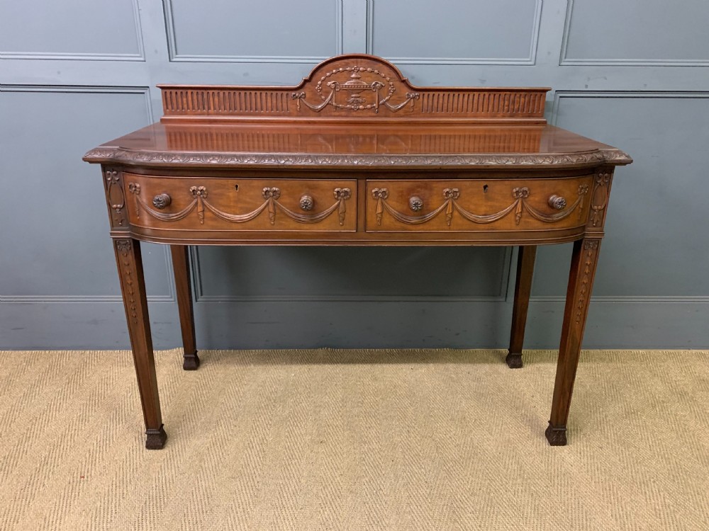 An Adam style mahogany side table featuring a carved upstand with an urn motif, relief drapery swags on the drawer fronts, and square fluted legs. View III