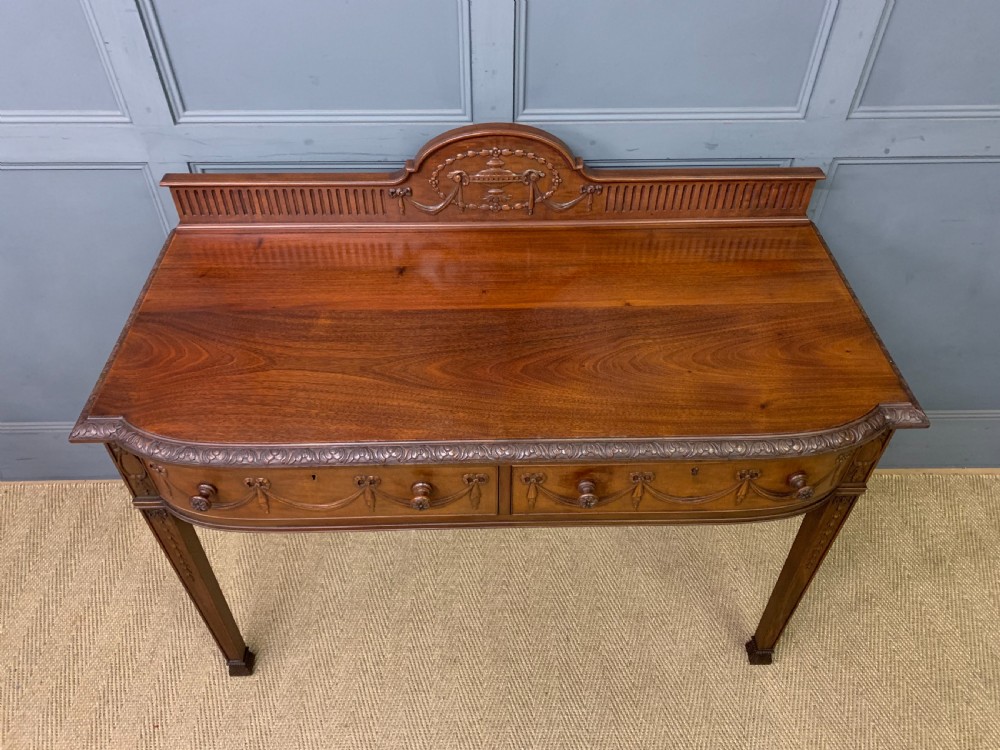 An Adam style mahogany side table featuring a carved upstand with an urn motif, relief drapery swags on the drawer fronts, and square fluted legs. View IV