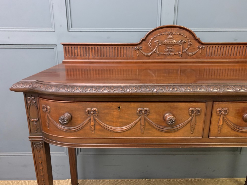 An Adam style mahogany side table featuring a carved upstand with an urn motif, relief drapery swags on the drawer fronts, and square fluted legs. View VII
