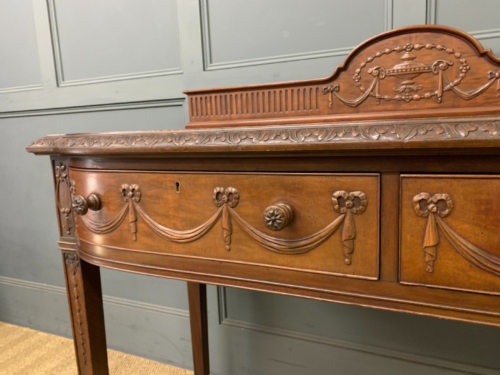 An Adam style mahogany side table featuring a carved upstand with an urn motif, relief drapery swags on the drawer fronts, and square fluted legs. View VIII