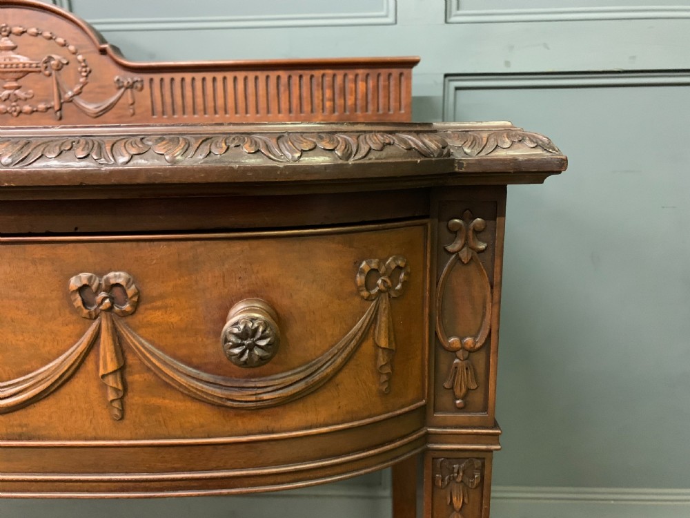 An Adam style mahogany side table featuring a carved upstand with an urn motif, relief drapery swags on the drawer fronts, and square fluted legs. View X