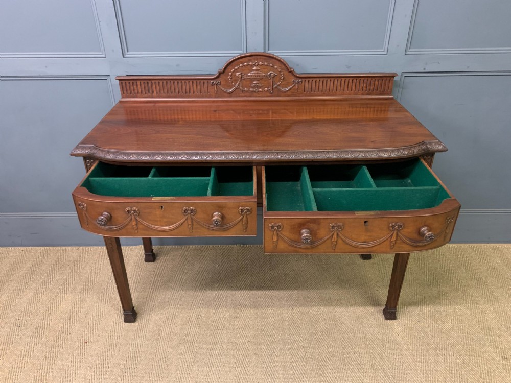 An Adam style mahogany side table featuring a carved upstand with an urn motif, relief drapery swags on the drawer fronts, and square fluted legs. View XII