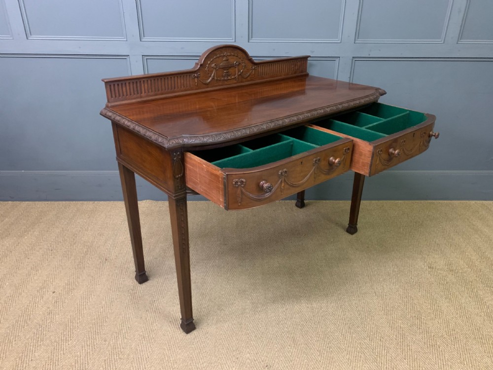 An Adam style mahogany side table featuring a carved upstand with an urn motif, relief drapery swags on the drawer fronts, and square fluted legs. View XVII