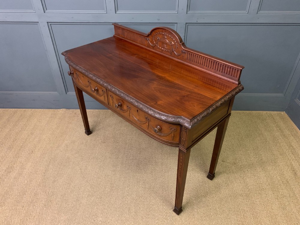 An Adam style mahogany side table featuring a carved upstand with an urn motif, relief drapery swags on the drawer fronts, and square fluted legs. View XVIII