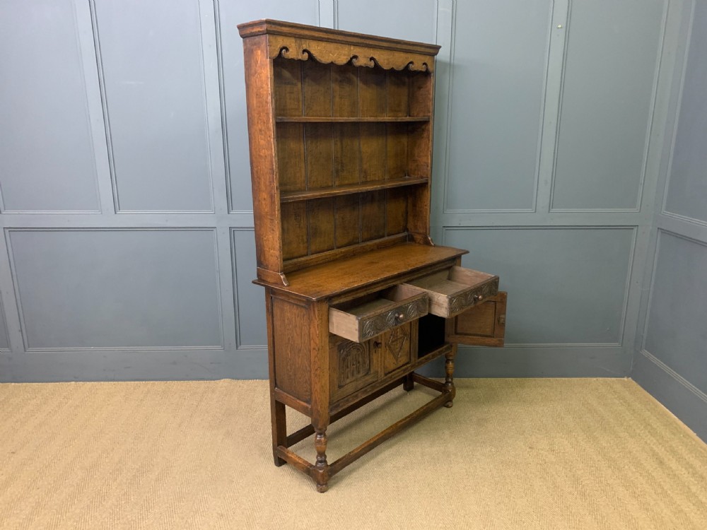 A carved oak Welsh dresser featuring an upper open shelving rack above two carved drawers and panelled cupboard doors resting on turned legs. View XVI