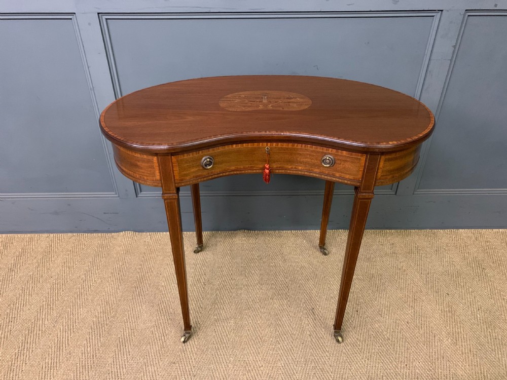 An Edwardian kidney-shaped mahogany table with central marquetry inlay, a single drawer with brass handles, and four tapered legs on castors.