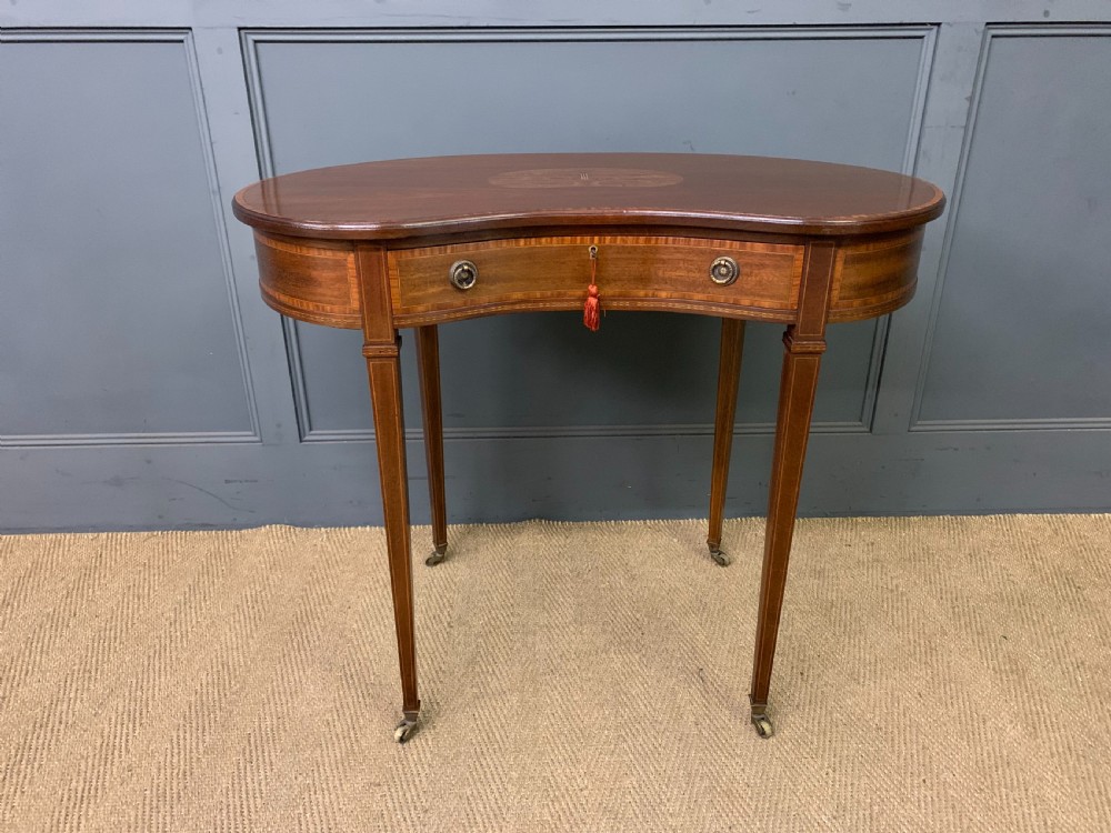 An Edwardian kidney-shaped mahogany table with central marquetry inlay, a single drawer with brass handles, and four tapered legs on castors. View IV
