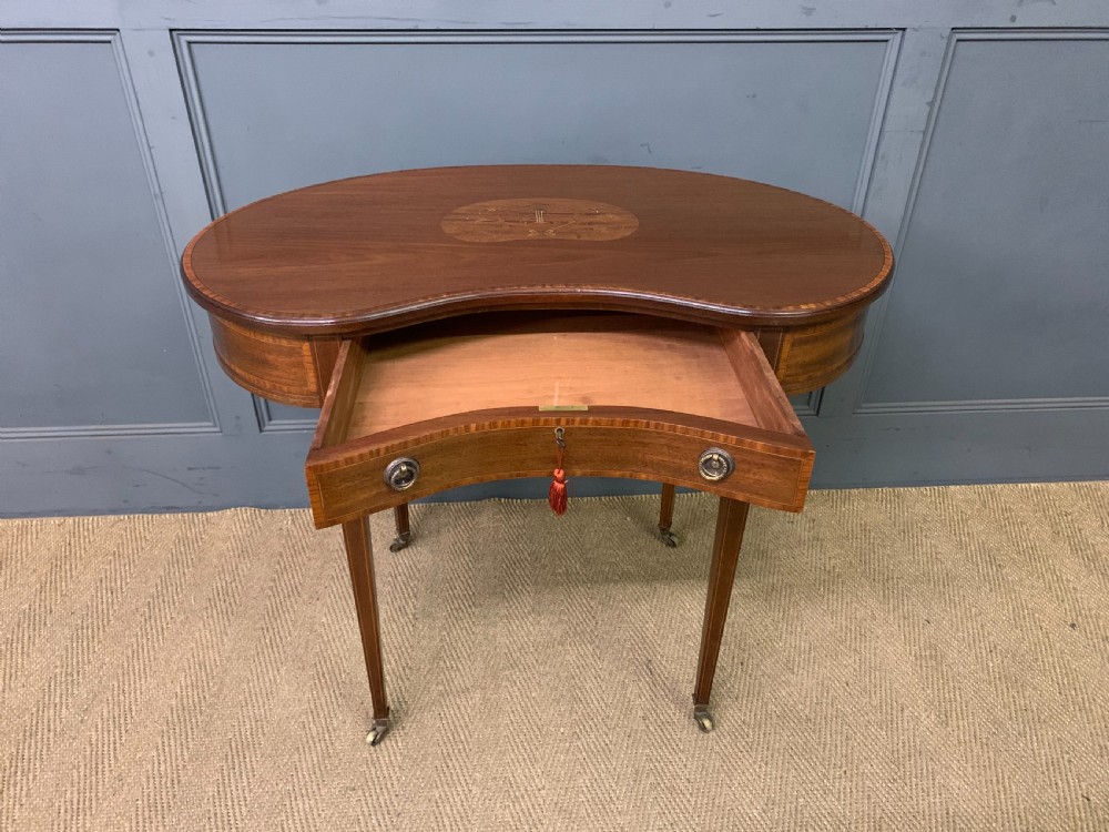 An Edwardian kidney-shaped mahogany table with central marquetry inlay, a single drawer with brass handles, and four tapered legs on castors. View V