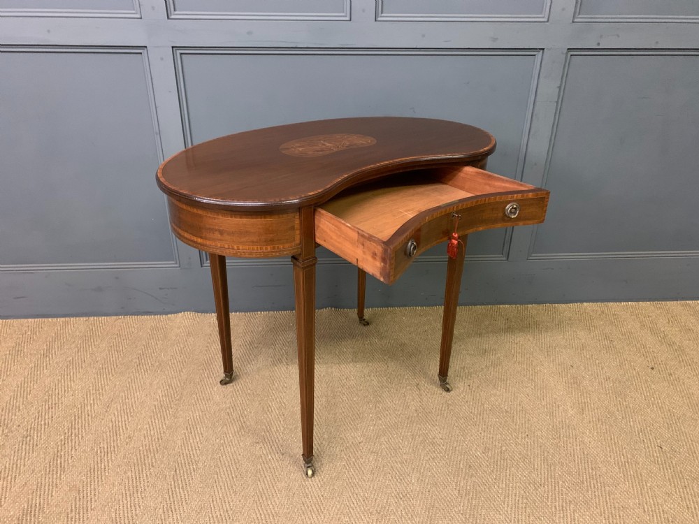 An Edwardian kidney-shaped mahogany table with central marquetry inlay, a single drawer with brass handles, and four tapered legs on castors. View VII