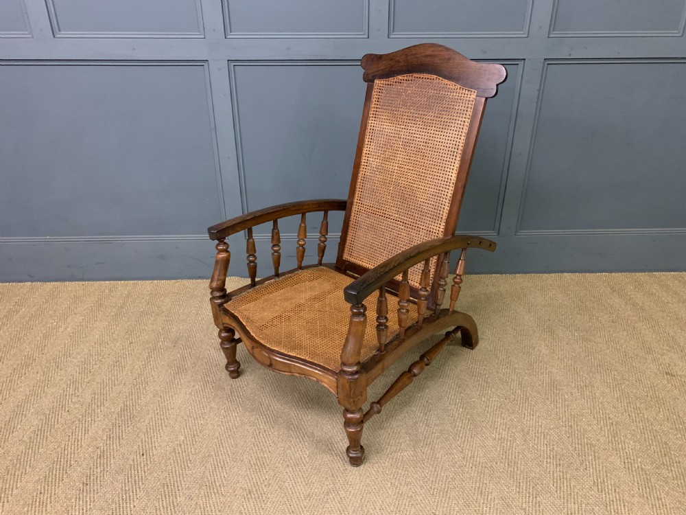 A 19th-century colonial reclining armchair crafted from dark wood with a hand-woven cane backrest and seat, featuring turned spindle arm supports.
