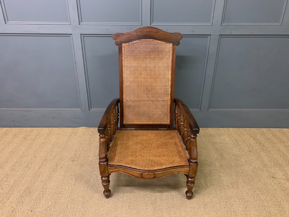 A 19th-century colonial reclining armchair crafted from dark wood with a hand-woven cane backrest and seat, featuring turned spindle arm supports. View II
