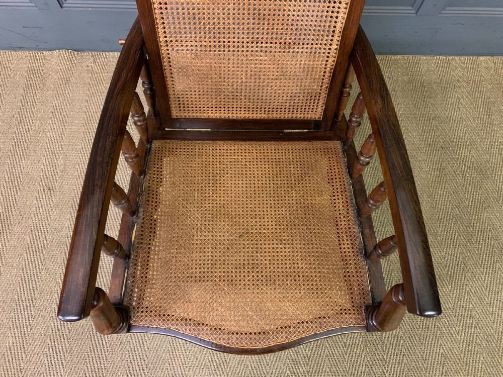 A 19th-century colonial reclining armchair crafted from dark wood with a hand-woven cane backrest and seat, featuring turned spindle arm supports. View V