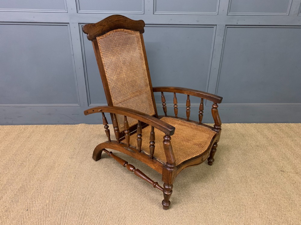 A 19th-century colonial reclining armchair crafted from dark wood with a hand-woven cane backrest and seat, featuring turned spindle arm supports. View VI