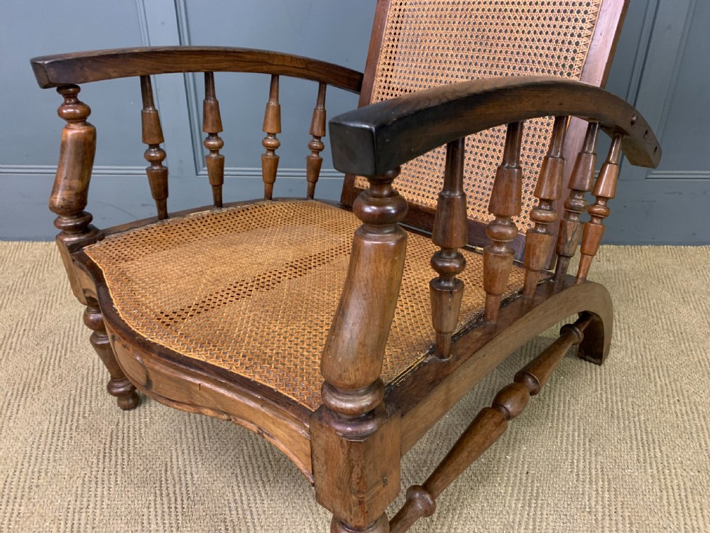 A 19th-century colonial reclining armchair crafted from dark wood with a hand-woven cane backrest and seat, featuring turned spindle arm supports. View IX