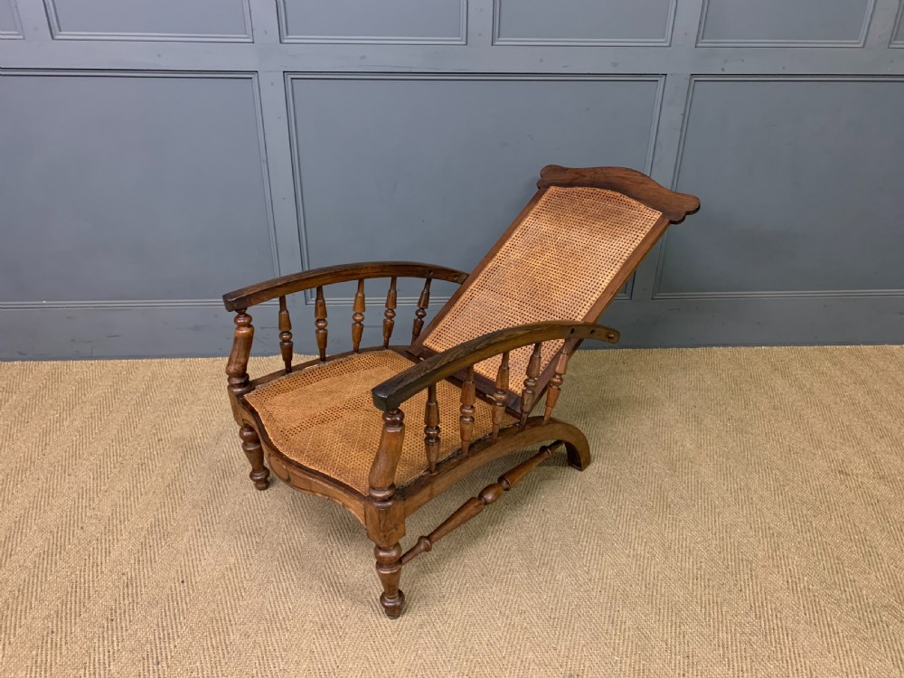 A 19th-century colonial reclining armchair crafted from dark wood with a hand-woven cane backrest and seat, featuring turned spindle arm supports. View X
