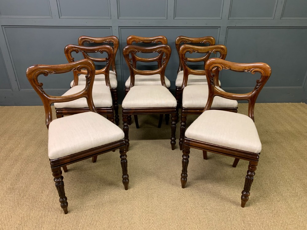 A set of eight early Victorian rosewood dining chairs featuring rounded balloon back frames, carved rail detailing, turned legs, and neutral upholstered seats.