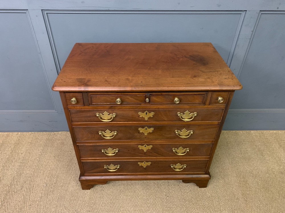 A Georgian mahogany bachelor chest featuring multiple drawers, brass handles, bracket feet, and an extended writing slide lined with green baize. View V