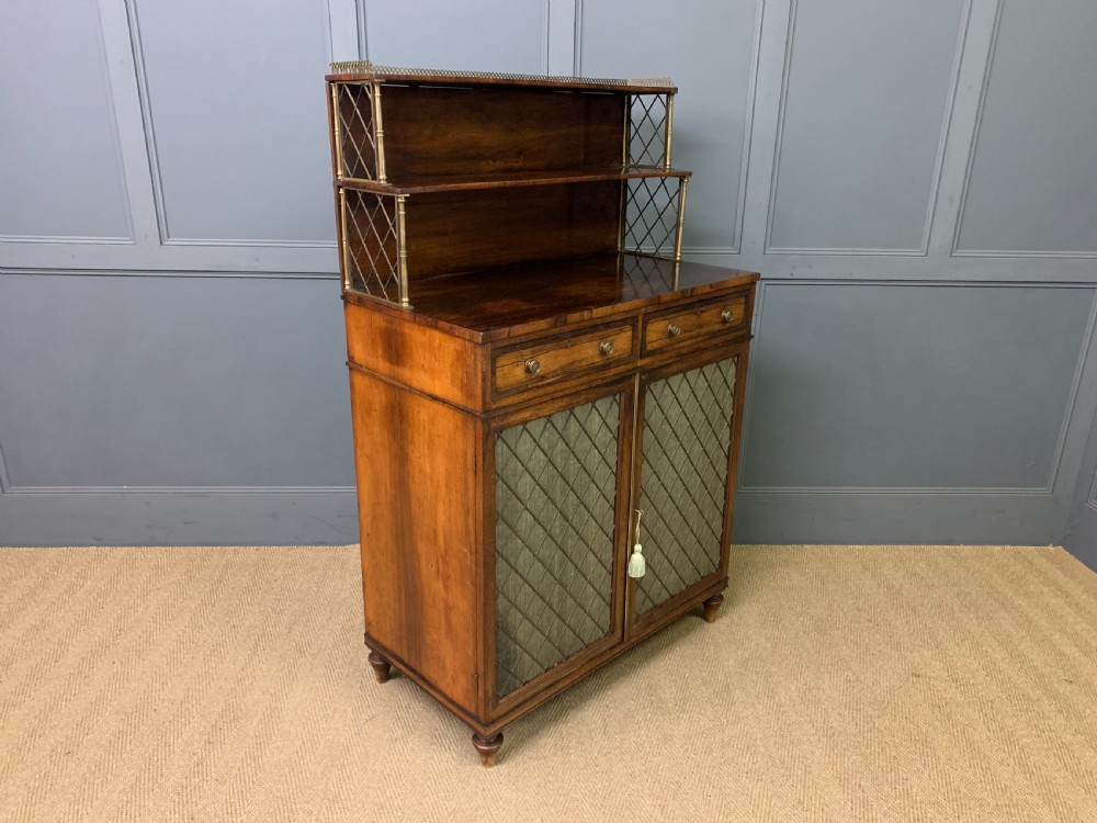 An antique Regency rosewood chiffonier featuring a brass lattice gallery shelf, two upper drawers, and lower doors with wire mesh and pleated fabric panels.