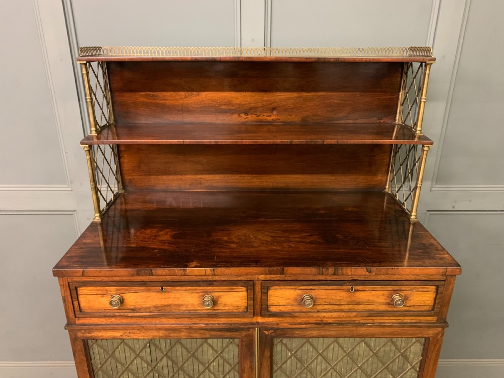 An antique Regency rosewood chiffonier featuring a brass lattice gallery shelf, two upper drawers, and lower doors with wire mesh and pleated fabric panels. View V