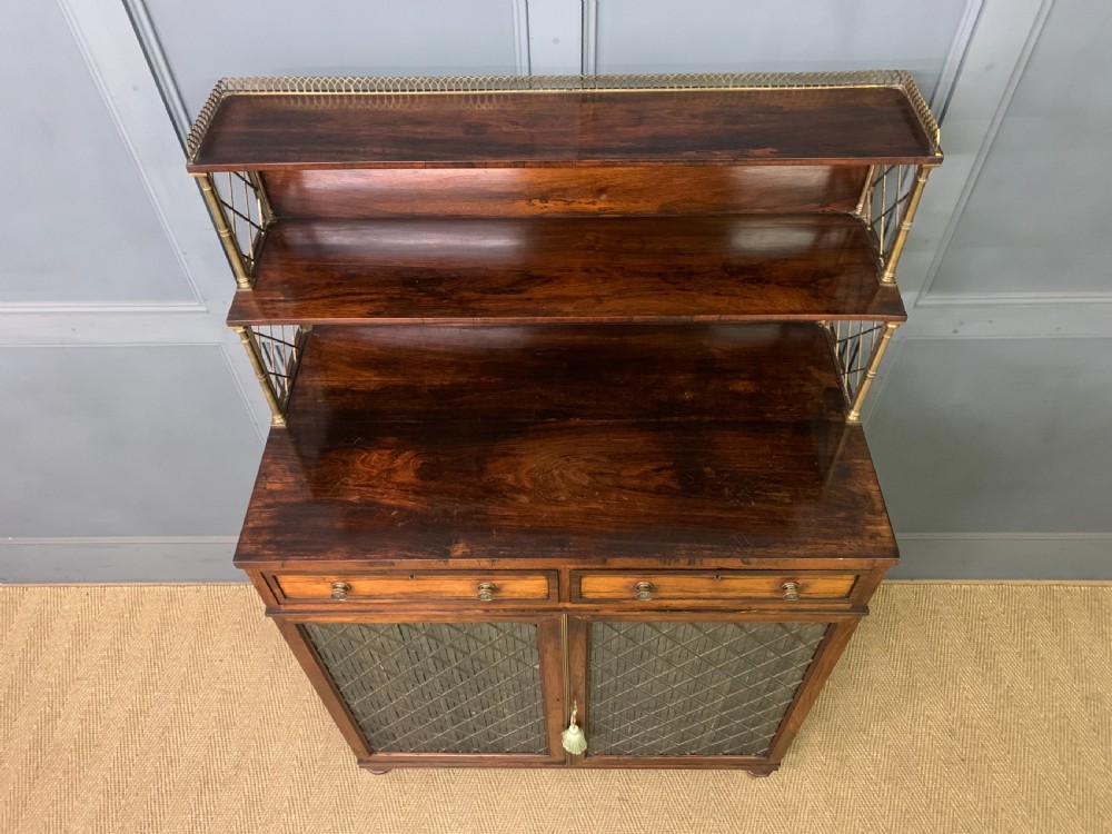 An antique Regency rosewood chiffonier featuring a brass lattice gallery shelf, two upper drawers, and lower doors with wire mesh and pleated fabric panels. View VI