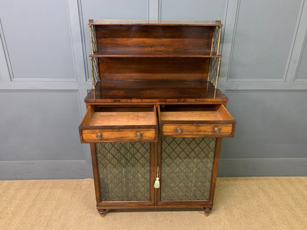 An antique Regency rosewood chiffonier featuring a brass lattice gallery shelf, two upper drawers, and lower doors with wire mesh and pleated fabric panels. View XIV