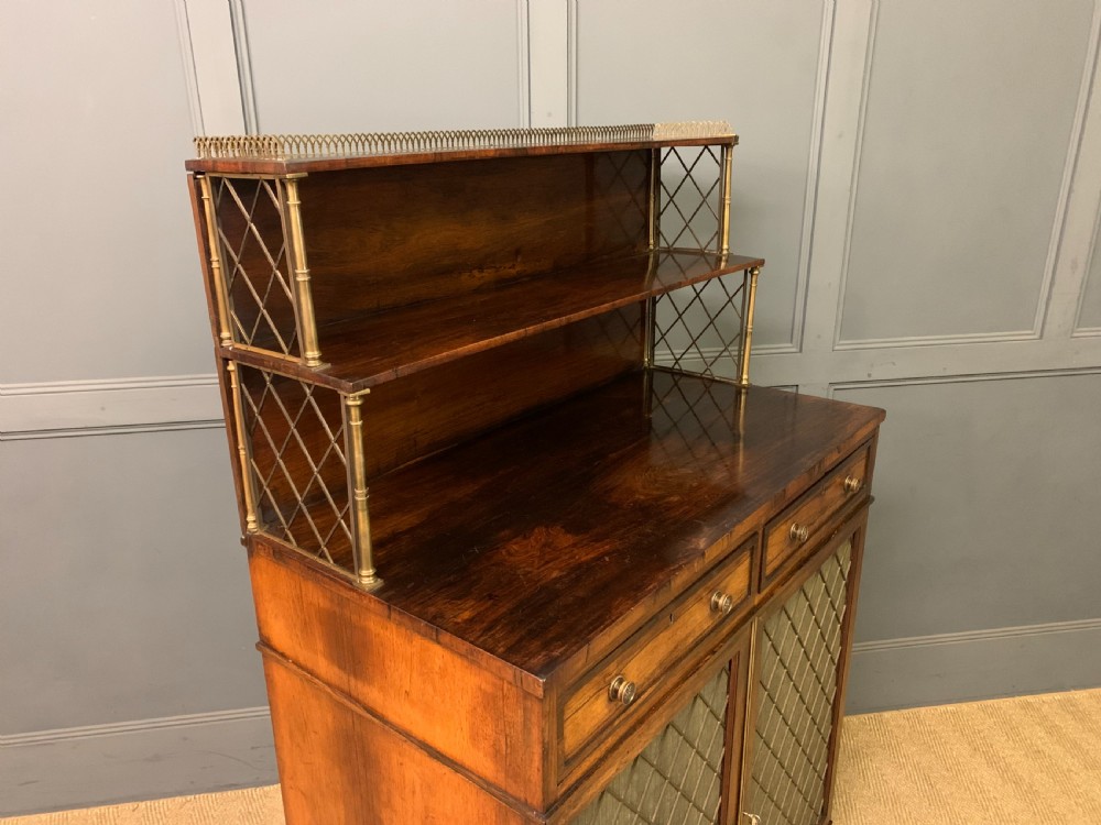 An antique Regency rosewood chiffonier featuring a brass lattice gallery shelf, two upper drawers, and lower doors with wire mesh and pleated fabric panels. View XVII