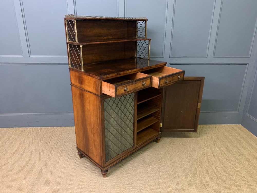 An antique Regency rosewood chiffonier featuring a brass lattice gallery shelf, two upper drawers, and lower doors with wire mesh and pleated fabric panels. View XIX