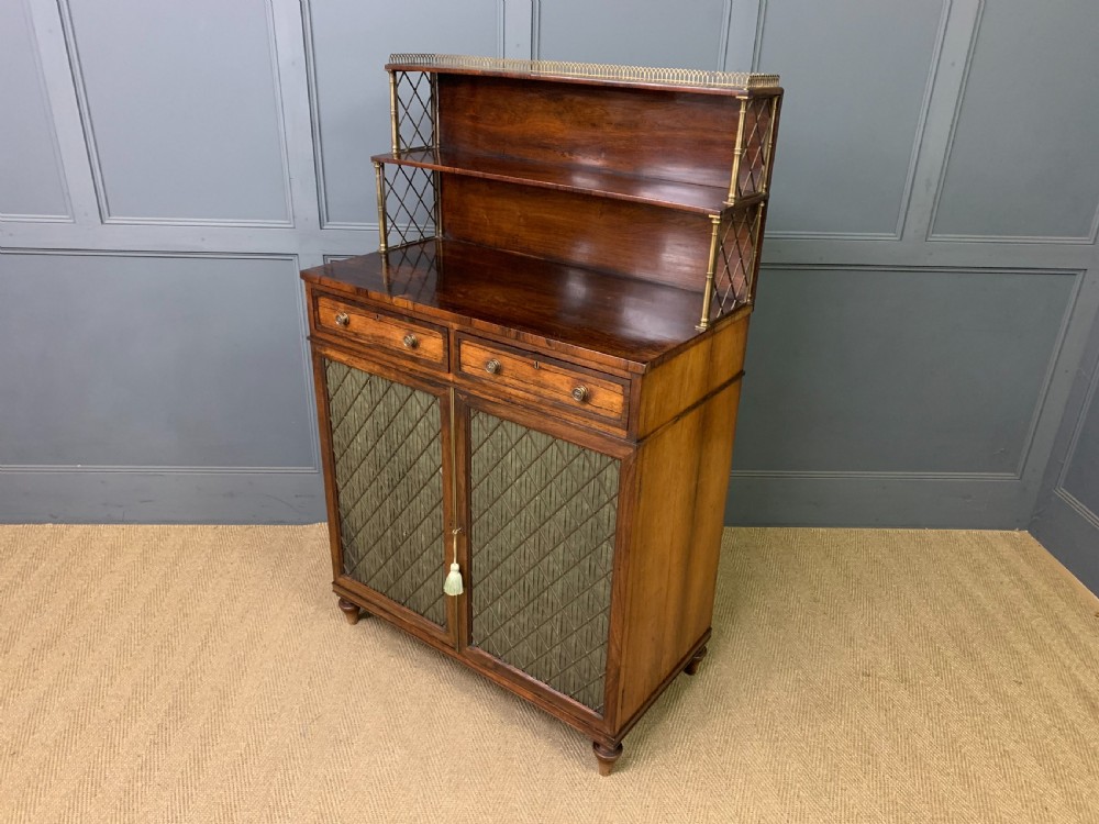 An antique Regency rosewood chiffonier featuring a brass lattice gallery shelf, two upper drawers, and lower doors with wire mesh and pleated fabric panels. View XX