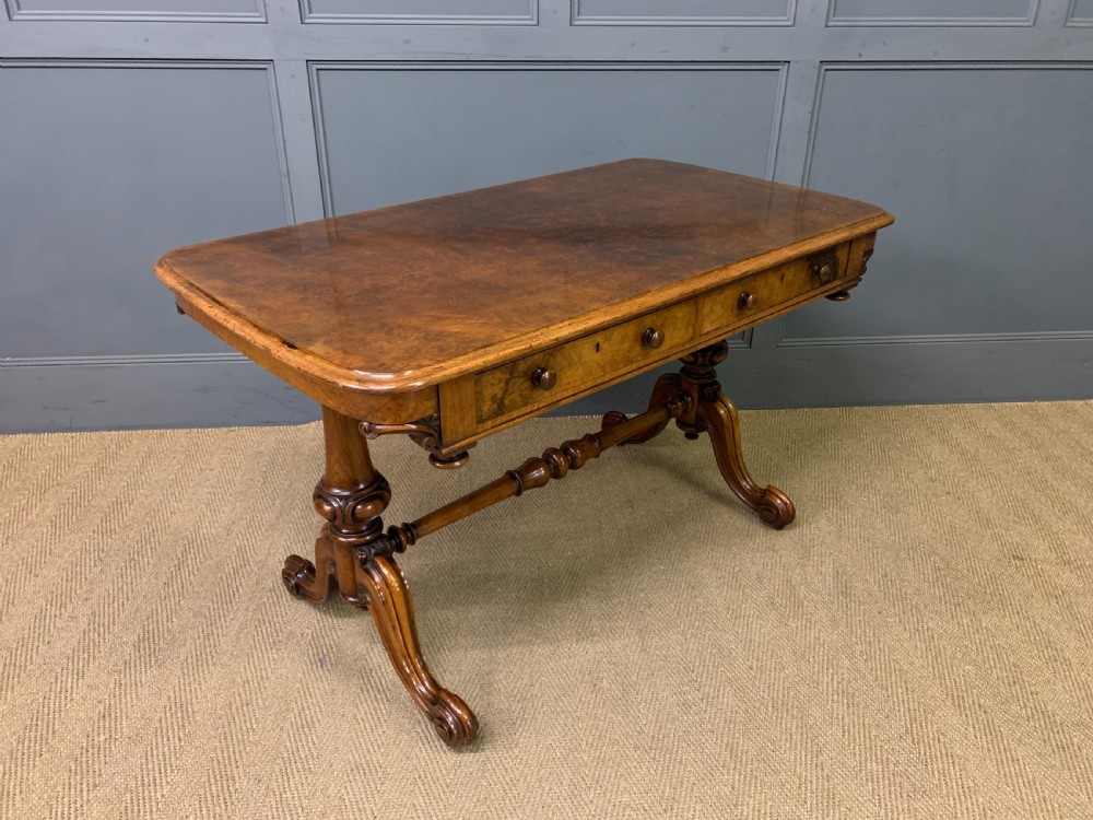 A Victorian burr walnut library table featuring a polished rectangular top with rounded corners, frieze drawers with wooden turned knobs, and twin carved end supports joined by a turned stretcher.
