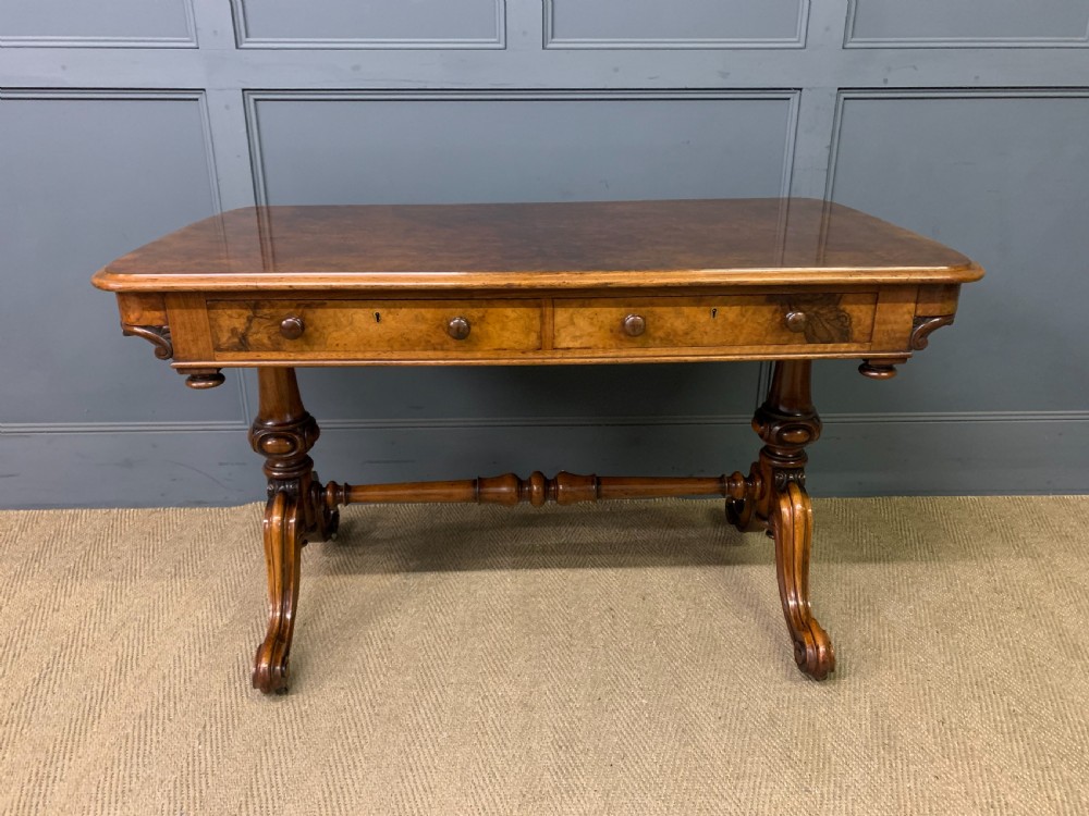 A Victorian burr walnut library table featuring a polished rectangular top with rounded corners, frieze drawers with wooden turned knobs, and twin carved end supports joined by a turned stretcher. View III