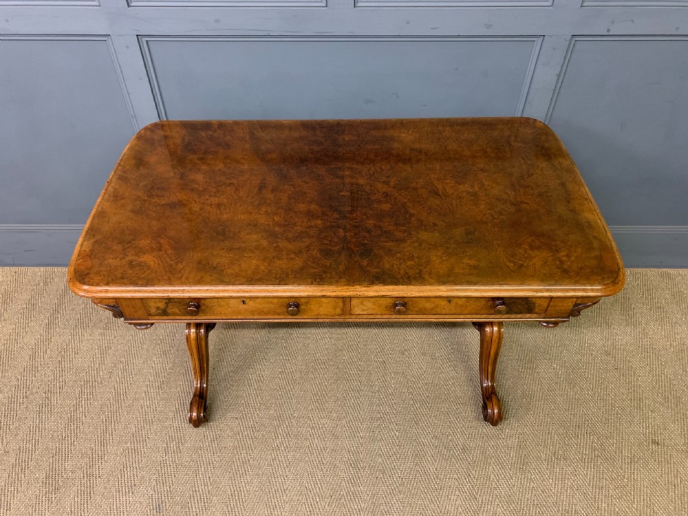 A Victorian burr walnut library table featuring a polished rectangular top with rounded corners, frieze drawers with wooden turned knobs, and twin carved end supports joined by a turned stretcher. View V