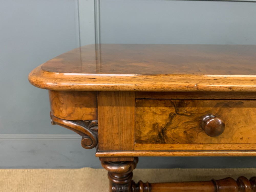 A Victorian burr walnut library table featuring a polished rectangular top with rounded corners, frieze drawers with wooden turned knobs, and twin carved end supports joined by a turned stretcher. View VI